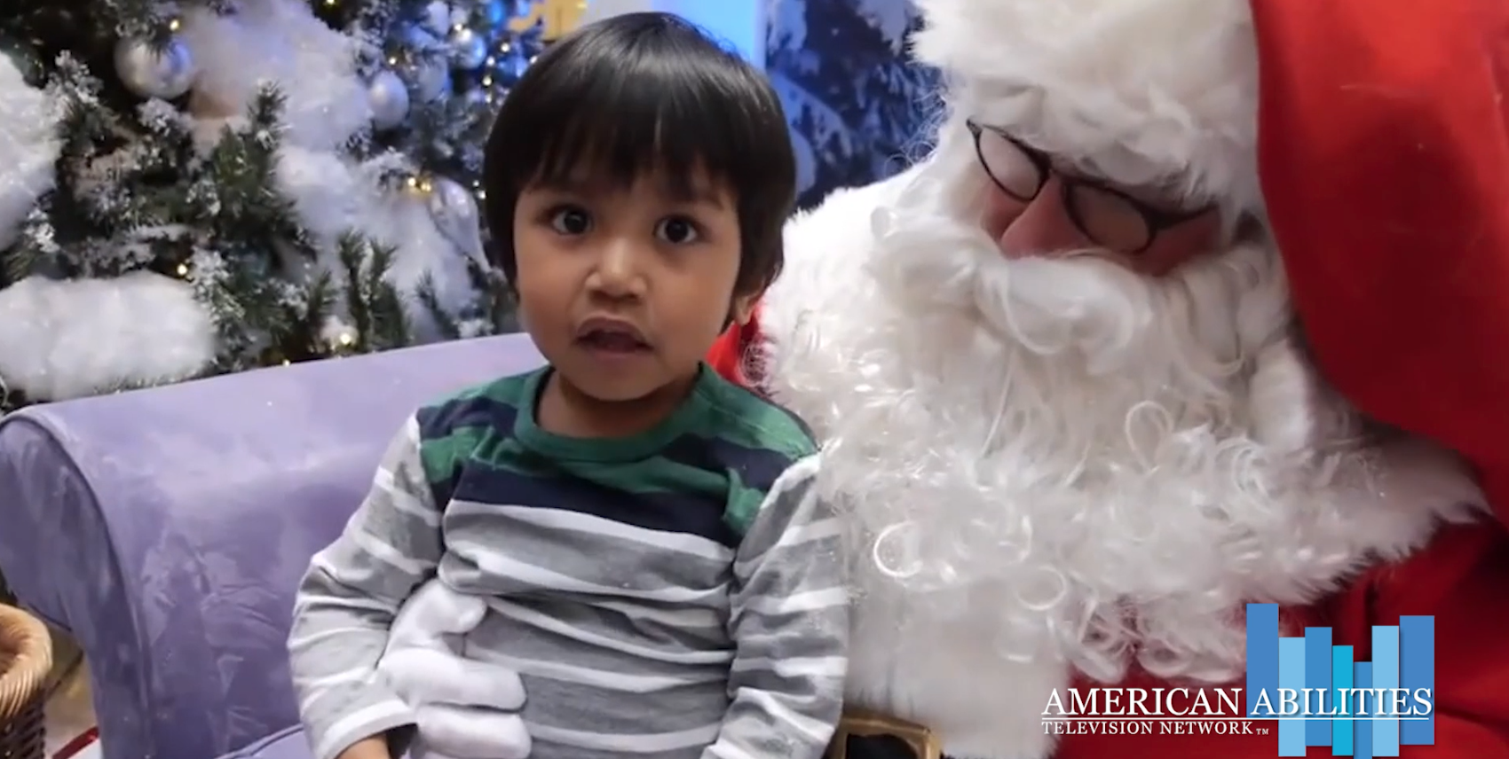 A young child with wide, surprised eyes seated on Santa Claus's lap in front of a frosted Christmas tree, in a still from the Santa Brings Christmas Joy To Deaf Children segment