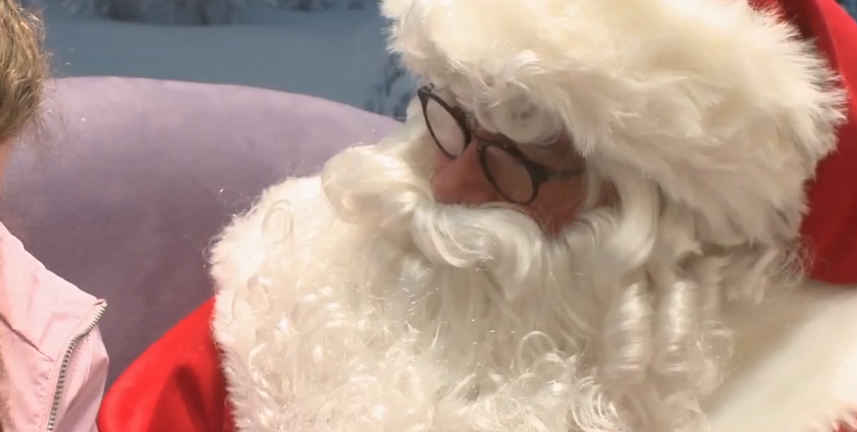 Santa Claus in round dark-rimmed glasses looking down toward a child in a pink jacket, in a still from the Santa Brings Christmas Joy To Deaf Children segment