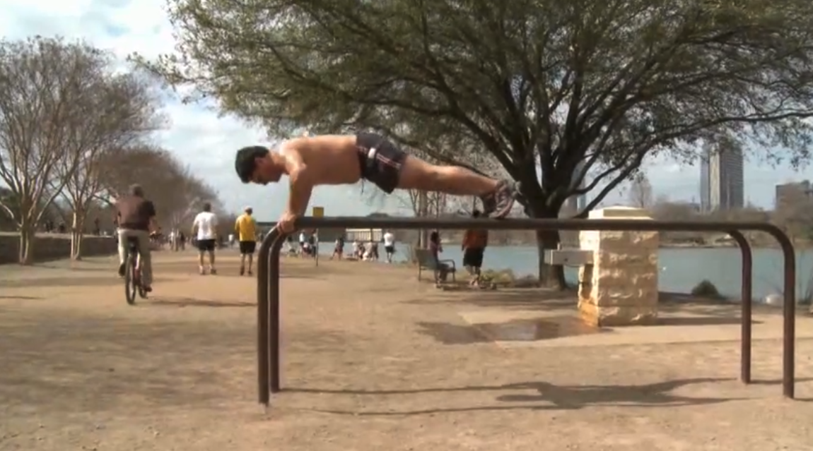 A man doing push-ups on outdoor exercise bars beside a riverfront trail, with cyclists and joggers passing, in a still from the CDC film A National Movement to Reduce Obesity and Tobacco Use