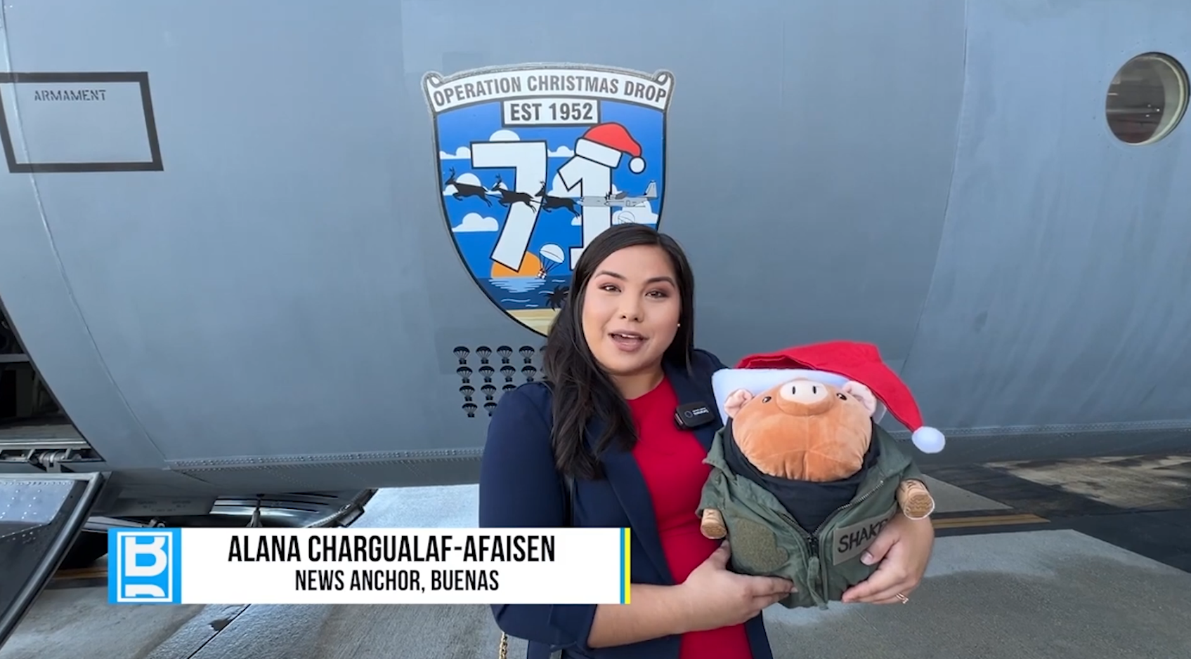 Buenas news anchor Alana Chargualaf-Afaisen holding a plush toy in a flight jacket and Santa hat in front of the Operation Christmas Drop aircraft emblem, in a still from the Operation Christmas Drop segment