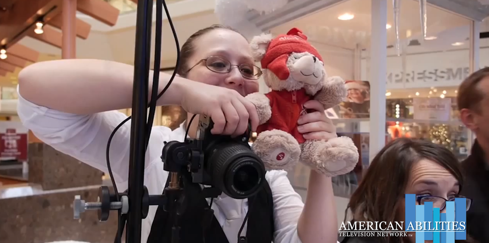 A photographer in a vest working a camera mounted on a stand while holding up a teddy bear dressed in a red hat and jacket to hold a child's attention, in a still from the Santa Brings Christmas Joy To Deaf Children segment
