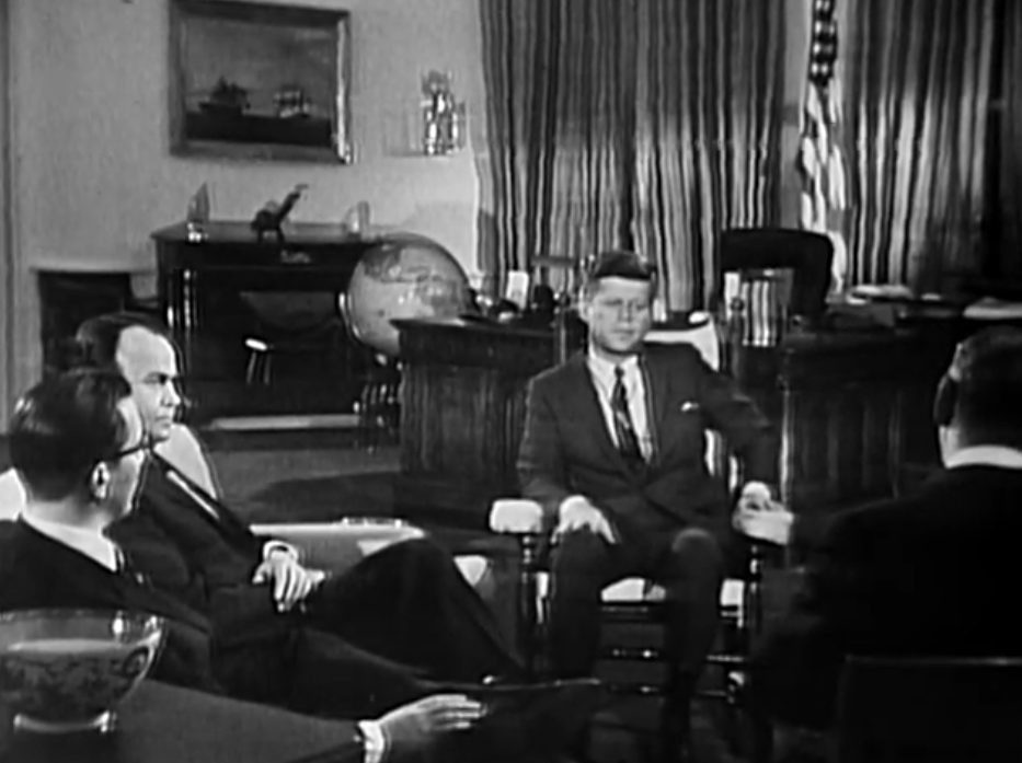 A second wide shot of the Oval Office during the 1962 After Two Years interview, with President Kennedy seated among the three correspondents and the desk, globe and flags visible, in a public-domain still from the original broadcast