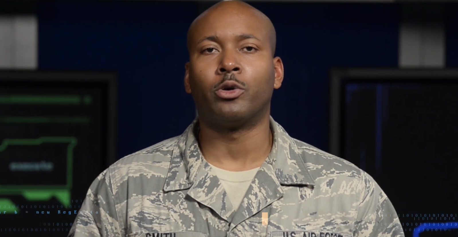 An airman with a Smith name tape and U.S. Air Force uniform speaking in a cyber studio with code graphics behind him, in a still from the U.S. Air Force BLUE series film The Cyber Shield